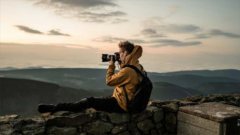 Hiker holding a compact outdoor camera while overlooking a misty mountain trail at sunrise